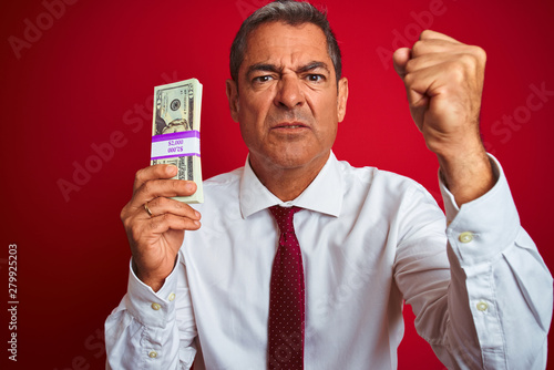 Handsome middle age businessman holding dollars over isolated red background annoyed and frustrated shouting with anger, crazy and yelling with raised hand, anger concept