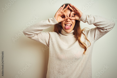 Young beautiful woman wearing winter sweater standing over white isolated background doing ok gesture like binoculars sticking tongue out, eyes looking through fingers. Crazy expression.