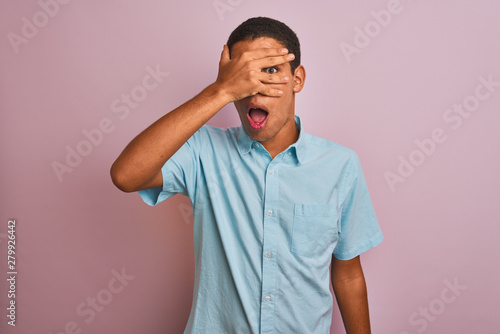 Young handsome arab man wearing blue shirt standing over isolated pink background peeking in shock covering face and eyes with hand, looking through fingers with embarrassed expression.
