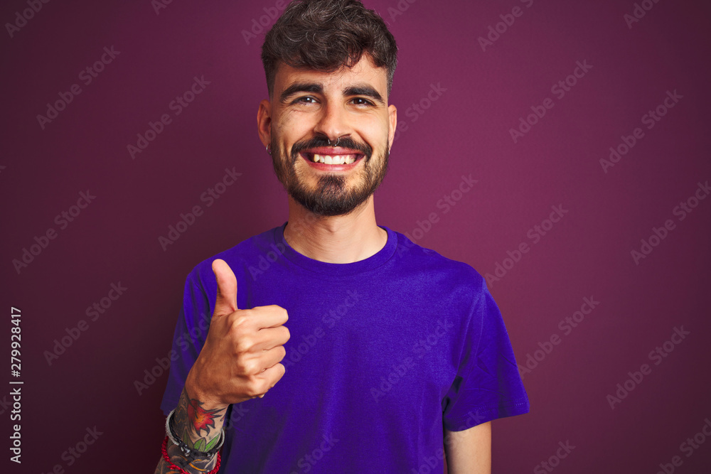Young man with tattoo wearing t-shirt standing over isolated purple background doing happy thumbs up gesture with hand. Approving expression looking at the camera showing success. Young man with tattoo wearing t-shirt standing over isolated purple background doing happy thumbs up gesture with hand. Approving expression looking at the camera showing success.