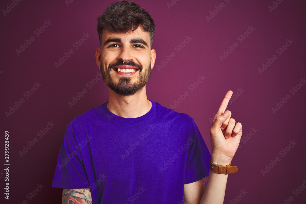 Young man with tattoo wearing t-shirt standing over isolated purple background with a big smile on face, pointing with hand and finger to the side looking at the camera. Young man with tattoo wearing t-shirt standing over isolated purple background with a big smile on face, pointing with hand and finger to the side looking at the camera.