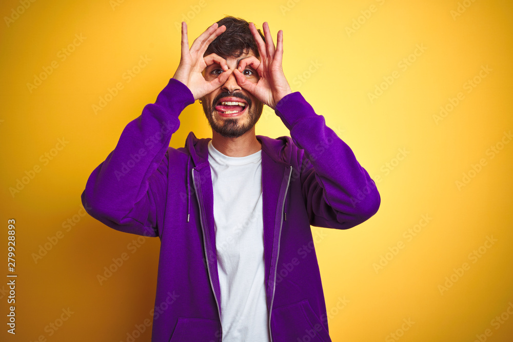 Young man with tattoo wearing sport purple sweatshirt over isolated yellow background doing ok gesture like binoculars sticking tongue out, eyes looking through fingers. Crazy expression. Young man with tattoo wearing sport purple sweatshirt over isolated yellow background doing ok gesture like binoculars sticking tongue out, eyes looking through fingers. Crazy expression.