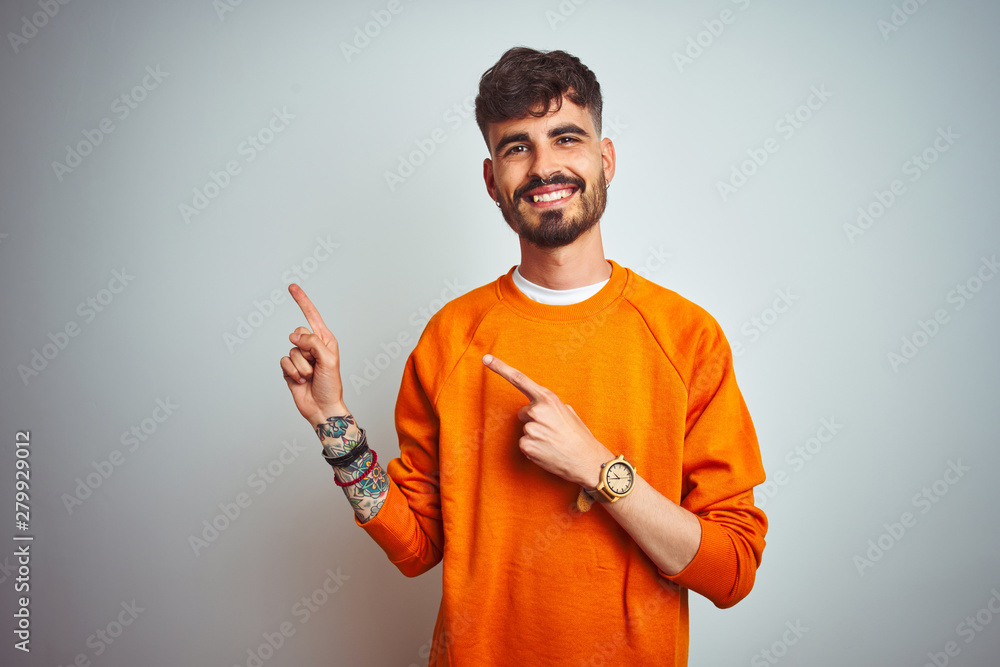 Young man with tattoo wearing orange sweater standing over isolated white background smiling and looking at the camera pointing with two hands and fingers to the side. Young man with tattoo wearing orange sweater standing over isolated white background smiling and looking at the camera pointing with two hands and fingers to the side.