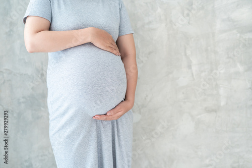 Asian pregnant woman in grey prenant dress holds hands on her big belly close up. Concept of healthy pregnant woman, prenant woman's portrait on concrete wall with copy space. photo