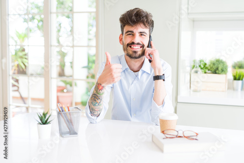 Young business man talking on smartphone happy with big smile doing ok sign, thumb up with fingers, excellent sign