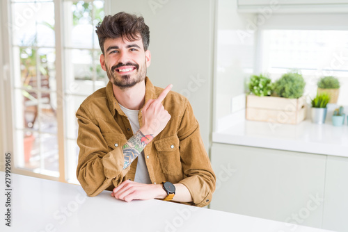 Young man wearing casual jacket sitting on white table cheerful with a smile of face pointing with hand and finger up to the side with happy and natural expression on face