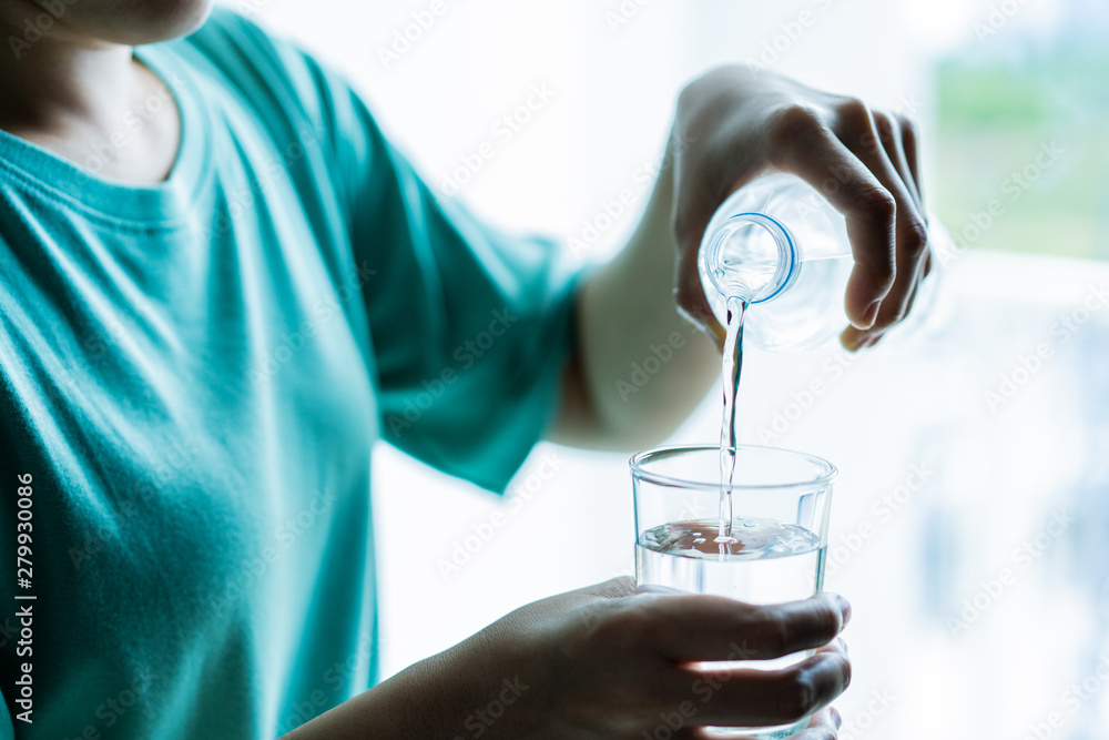 Woman pouring a mineral drinking water from a plastic water bottle into glass close up. Woman pouring mineral water into glass near the window. Concept of health care and good health life style. Woman pouring a mineral drinking water from a plastic water bottle into glass close up. Woman pouring mineral water into glass near the window. Concept of health care and good health life style.