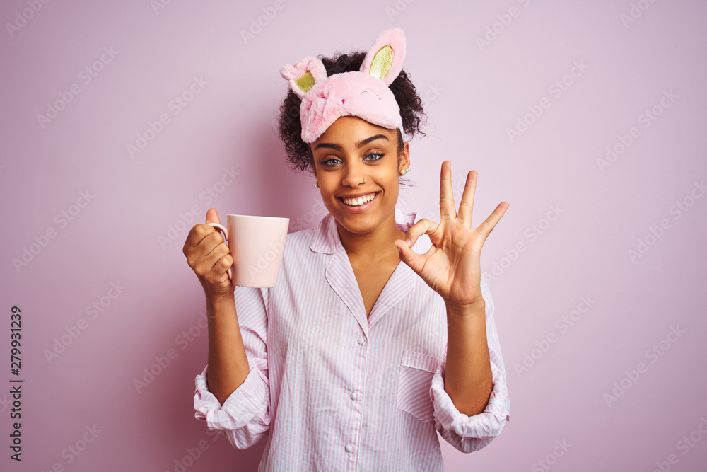 Afro woman wearing pajama and mask drinking a cup of coffee over isolated pink background doing ok sign with fingers, excellent symbol - obrazy, fototapety, plakaty 
