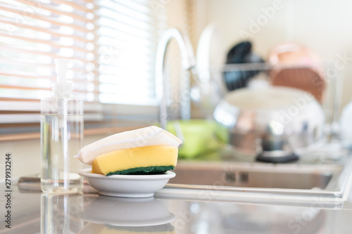 Yellow sponges and dish washing liquid soap on dirty sink fully with dishes and kitchen ware.
