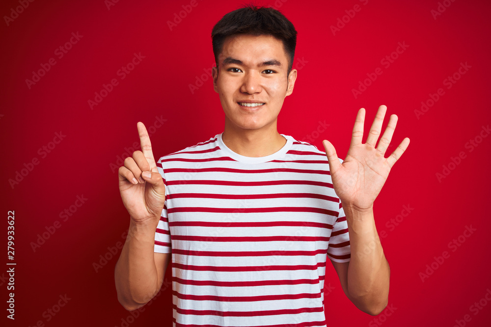 Young asian chinese man wearing striped t-shirt standing over isolated red background showing and pointing up with fingers number six while smiling confident and happy.