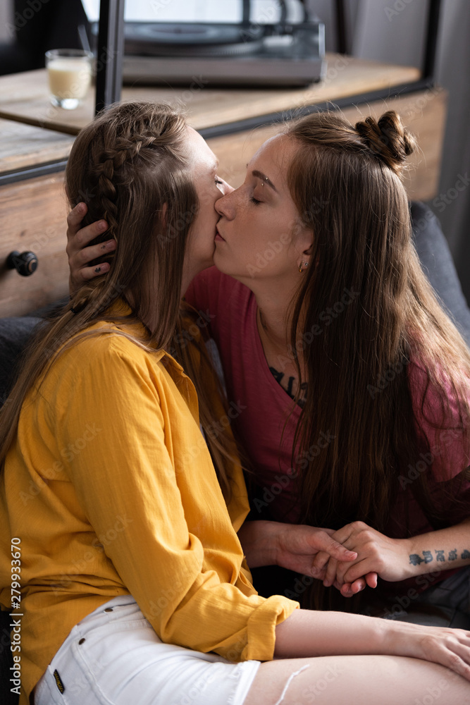 two lesbians kissing and holding hands while sitting on sofa in living room two lesbians kissing and holding hands while sitting on sofa in living room