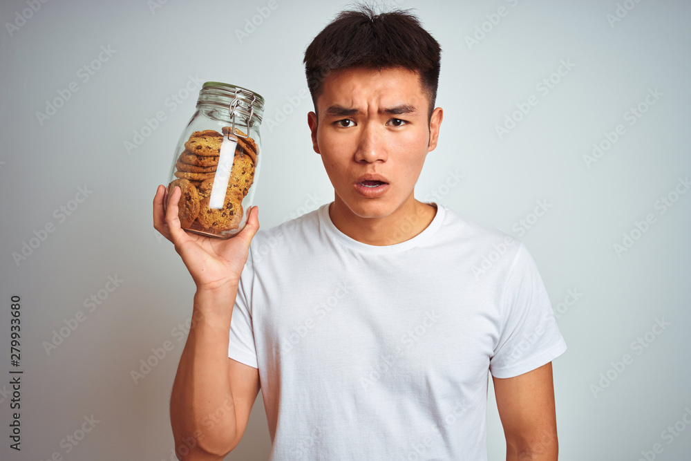 Young asian chinese man holding jar of cookies standing over isolated white background scared in shock with a surprise face, afraid and excited with fear expression Young asian chinese man holding jar of cookies standing over isolated white background scared in shock with a surprise face, afraid and excited with fear expression