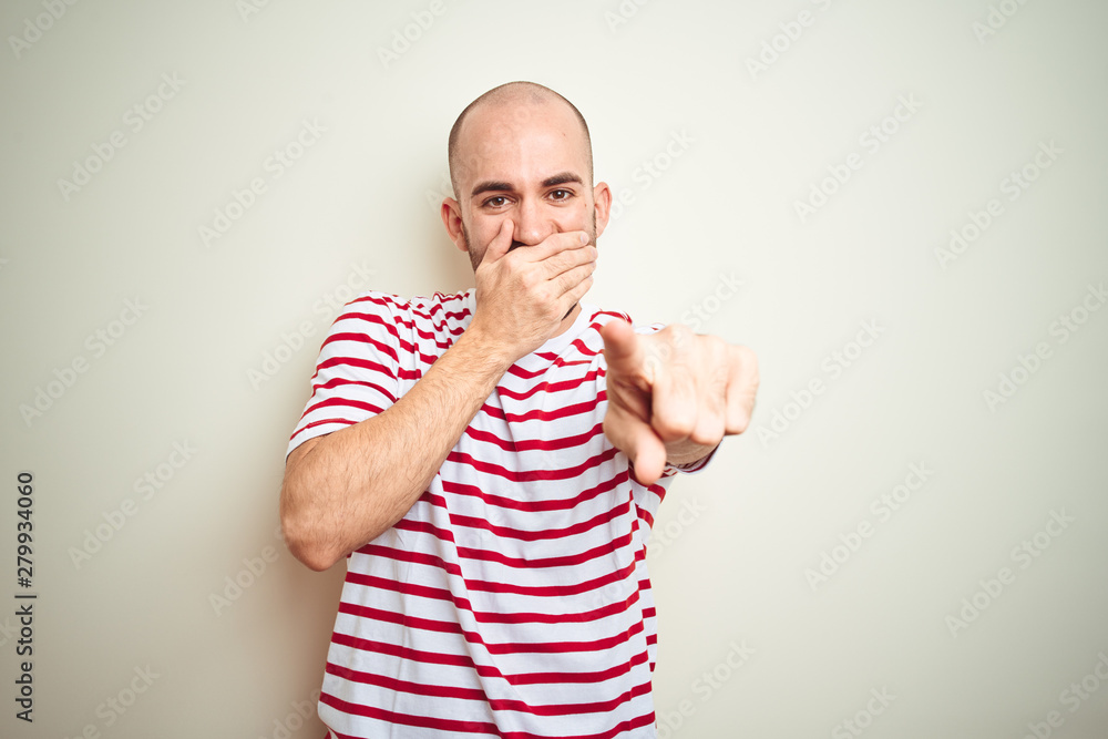 Young bald man with beard wearing casual striped red t-shirt over white isolated background laughing at you, pointing finger to the camera with hand over mouth, shame expression Young bald man with beard wearing casual striped red t-shirt over white isolated background laughing at you, pointing finger to the camera with hand over mouth, shame expression