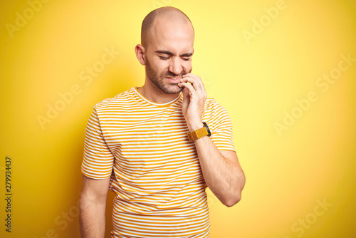 Young bald man with beard wearing casual striped t-shirt over yellow isolated background touching mouth with hand with painful expression because of toothache or dental illness on teeth. 