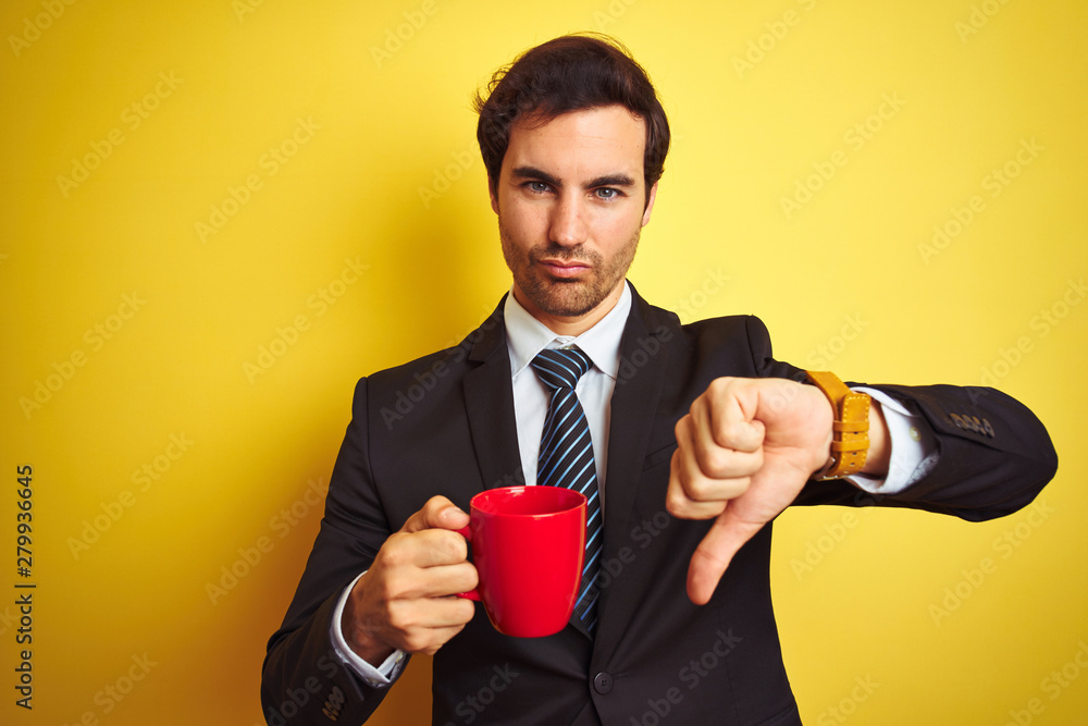 Young handsome businessman drinking red cup of coffee over isolated yellow background with angry face, negative sign showing dislike with thumbs down, rejection concept Young handsome businessman drinking red cup of coffee over isolated yellow background with angry face, negative sign showing dislike with thumbs down, rejection concept