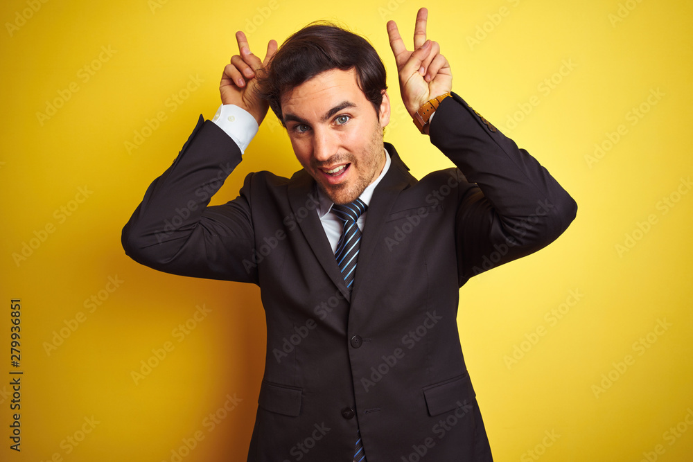 Young handsome businessman wearing suit and tie standing over isolated yellow background Posing funny and crazy with fingers on head as bunny ears, smiling cheerful Young handsome businessman wearing suit and tie standing over isolated yellow background Posing funny and crazy with fingers on head as bunny ears, smiling cheerful