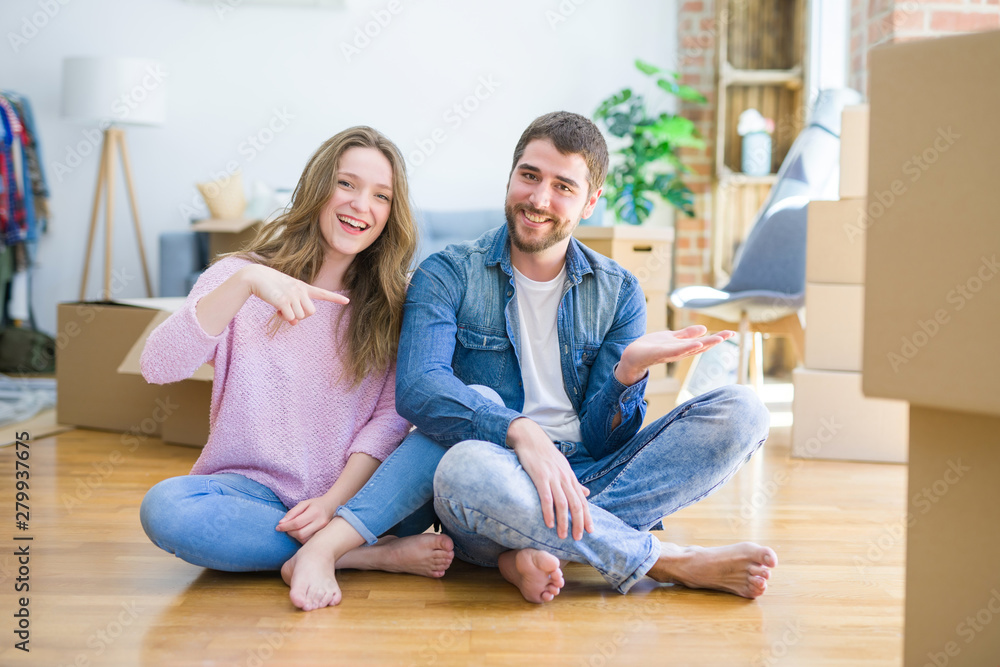 Young beautiful couple moving to a new house sitting on the floor amazed and smiling to the camera while presenting with hand and pointing with finger. Young beautiful couple moving to a new house sitting on the floor amazed and smiling to the camera while presenting with hand and pointing with finger.