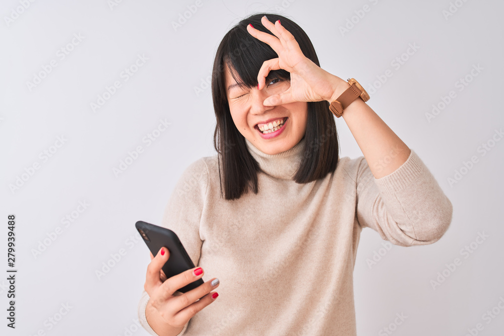 Young beautiful Chinese woman using smartphone standing over isolated white background with happy face smiling doing ok sign with hand on eye looking through fingers Young beautiful Chinese woman using smartphone standing over isolated white background with happy face smiling doing ok sign with hand on eye looking through fingers