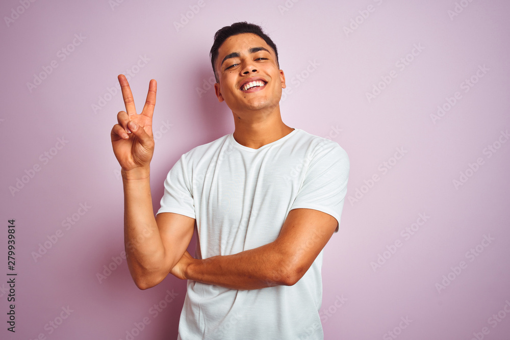 Young brazilian man wearing t-shirt standing over isolated pink background smiling with happy face winking at the camera doing victory sign. Number two. Young brazilian man wearing t-shirt standing over isolated pink background smiling with happy face winking at the camera doing victory sign. Number two.