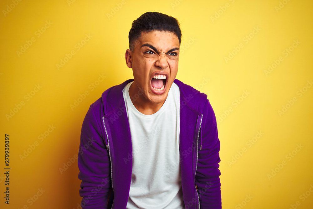 Young brazilian man wearing purple sweatshirt standing over isolated yellow background angry and mad screaming frustrated and furious, shouting with anger. Rage and aggressive concept. - obrazy, fototapety, plakaty 