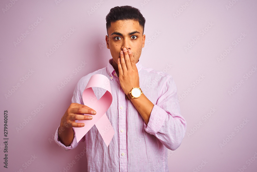 Young brazilian man holding cancer ribbon standing over isolated pink background cover mouth with hand shocked with shame for mistake, expression of fear, scared in silence, secret concept Young brazilian man holding cancer ribbon standing over isolated pink background cover mouth with hand shocked with shame for mistake, expression of fear, scared in silence, secret concept