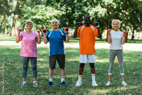cheerful multicultural pensioners in sportswear exercising with dumbbells in park