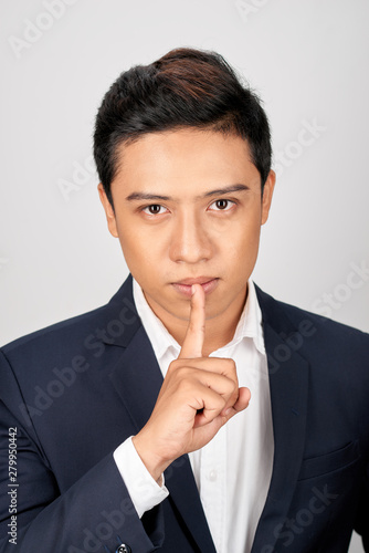 Portrait of Asian handsome businessman dressed in suit showing silence gesture and looking at camera isolated over white background