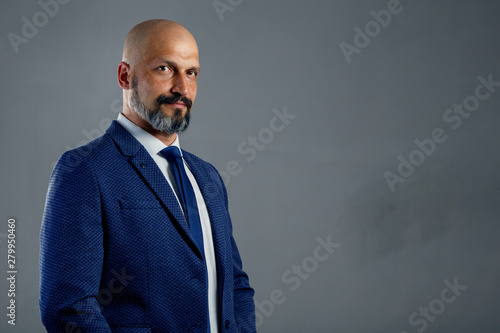 Brutal bald bearded businessman in a classic suit on a gray background with copy space
