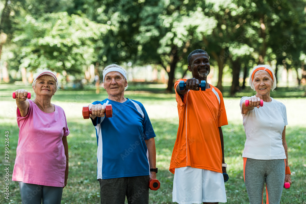 smiling multicultural pensioners exercising with dumbbells in park