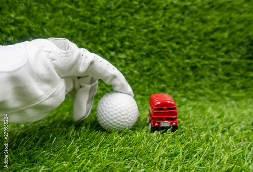 hand is pointing golf ball with red double decker bus are on green grass.