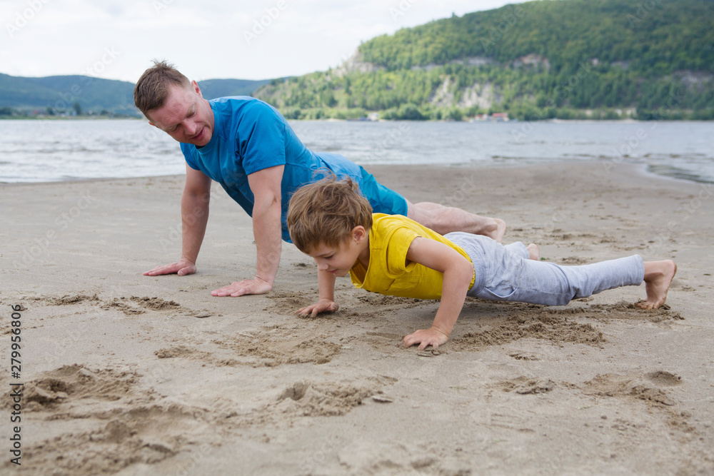 Father and son spend time together and lead a healthy lifestyle outdoors. Man and boy doing push ups are working out. Father and son are doing exercise on the beach in the summer. Father and son spend time together and lead a healthy lifestyle outdoors. Man and boy doing push ups are working out. Father and son are doing exercise on the beach in the summer.