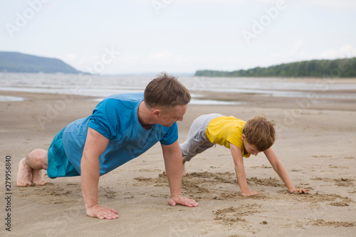  Father and son spend time together and lead a healthy lifestyle outdoors. Man and boy doing push ups are working out. Father and son are doing exercise on the beach in the summer.