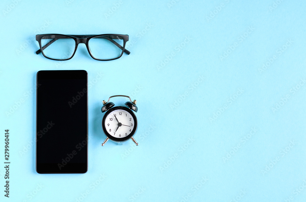 Black glasses, alarm clock and cellphone on blue background composition. Black glasses, alarm clock and cellphone on blue background composition.