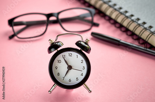 Black glasses, alarm clock and paper notepad on pink background composition.