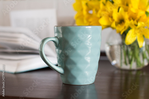 blue coffee mug, yellow flowers in transparent bowl and a book on a wooden table