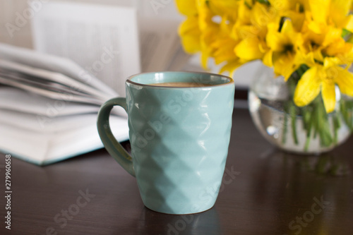 blue coffee mug, yellow flowers in transparent bowl and a book on a wooden table