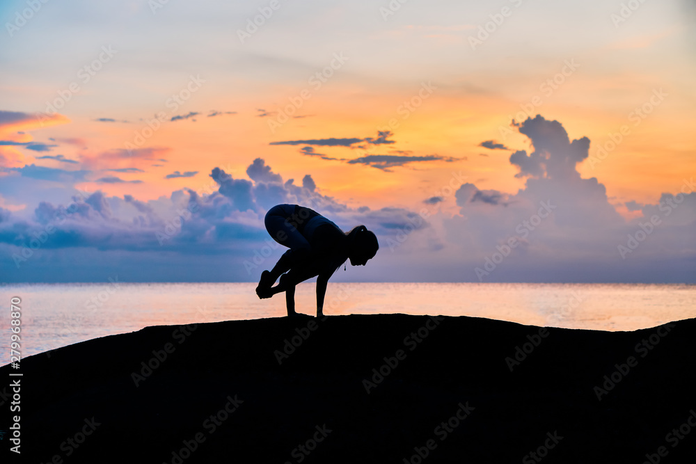 Silhouette of a young woman doing yoga exercises  by the sea against the backdrop of colourful dawn                  