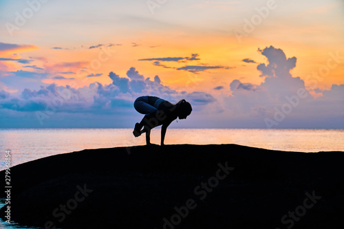 Silhouette of a young woman doing yoga exercises by the sea against the backdrop of colourful dawn 