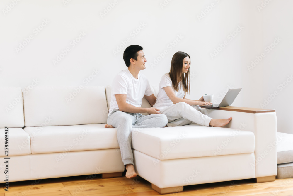 Young married couple sitting on soft sofa and looking at laptop in bright living room. Happy people buying things for house in online store sitting in light apartment. Guy and girl smiling. Young married couple sitting on soft sofa and looking at laptop in bright living room. Happy people buying things for house in online store sitting in light apartment. Guy and girl smiling.