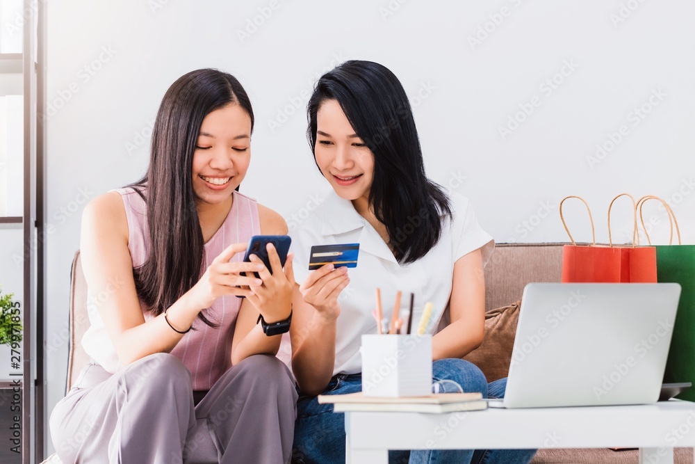 Asian beautiful woman and friend using smartphone with credit card shopping online together at home with shopping bag and laptop.Convenience buying online products with business technology concept. Asian beautiful woman and friend using smartphone with credit card shopping online together at home with shopping bag and laptop.Convenience buying online products with business technology concept.