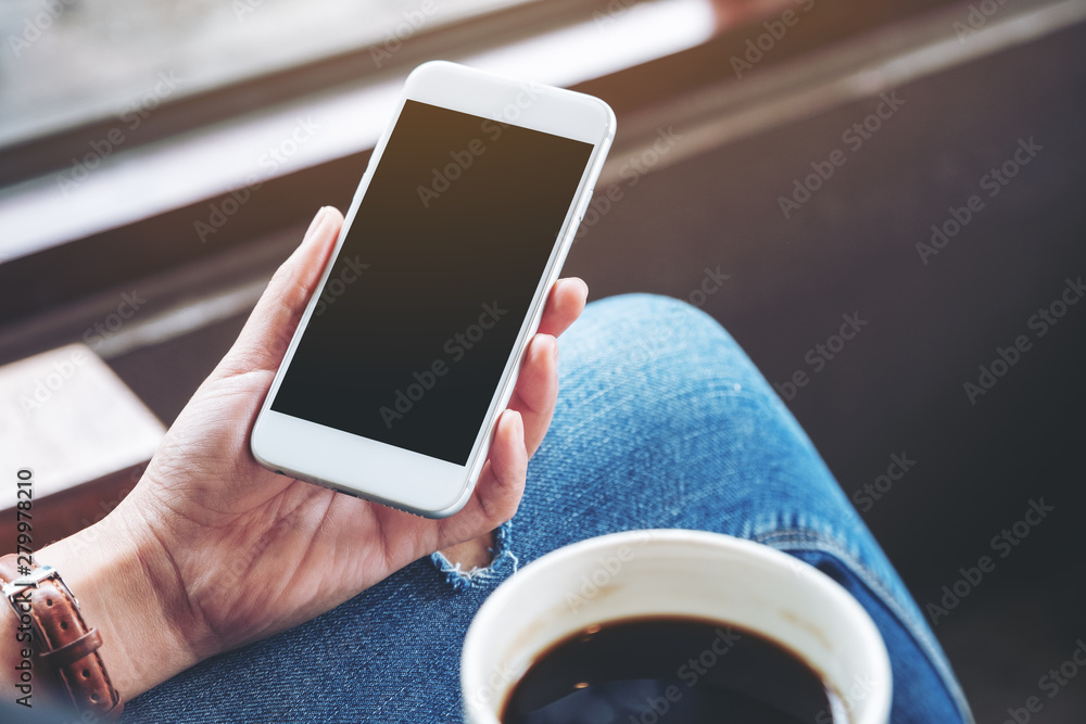 Mockup image of woman holding white mobile phone with blank desktop screen while drinking coffee in cafe