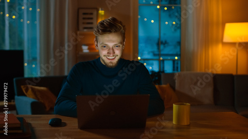 Handsome Smiling Man Works on a Laptop while Sitting at His Desk at Home. Young Freelancer Works on Computer in His Cozy Living Room with Warm Evening Lighting Turned on.