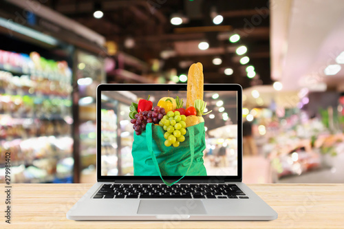 supermarket aisle blurred background with laptop computer and green shopping bag on wood table grocery online concept