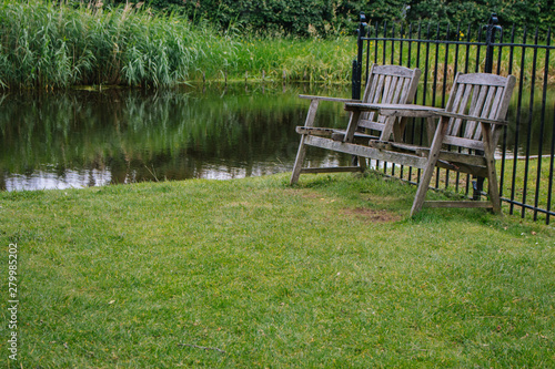 Two wooden chairs in the backyard near pond. Old outdoor furniture in summer garden. Empty quiet place for relax. Dutch patio with retro wooden bench. Lake with lawn and trees. Summer landscape. © Nataliia
