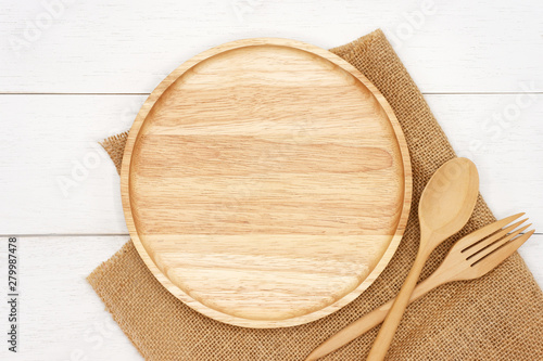 Empty round wooden plate with spoon, fork and rustic brown burlap cloth on white wooden table. Top view image.