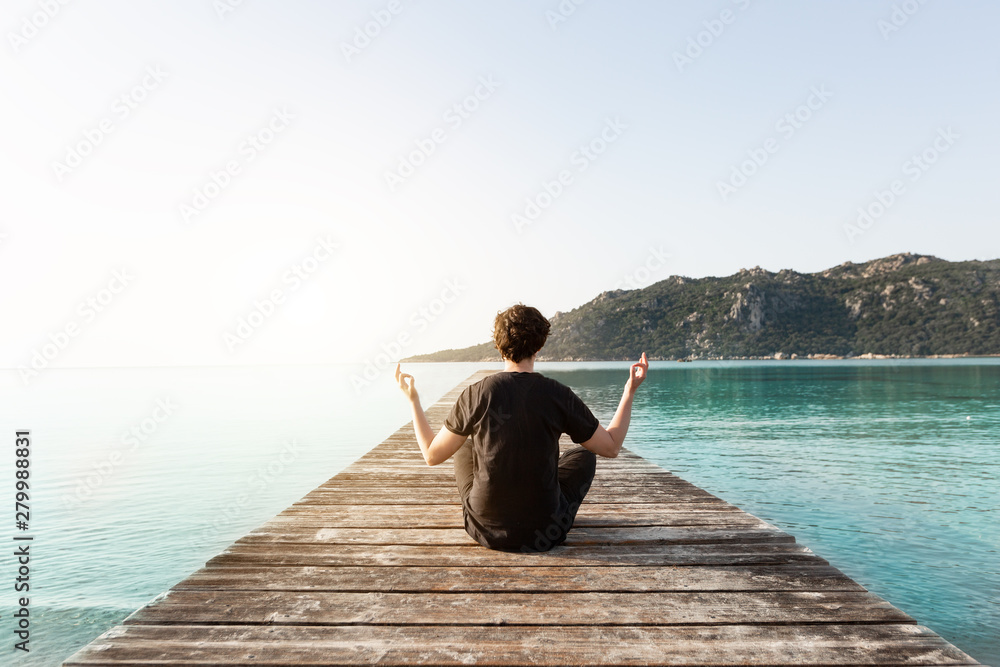 Person sitzt auf einem Steg und meditiert umgeben vom Meer beim Sonnenaufgang