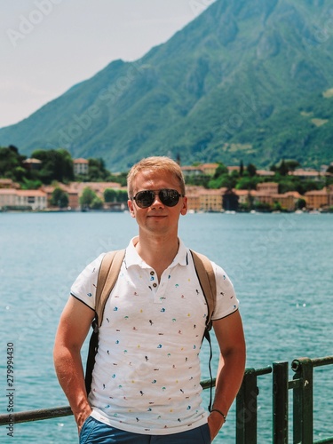A young blond man with a backpack on his shoulders stands with a view of lake Como