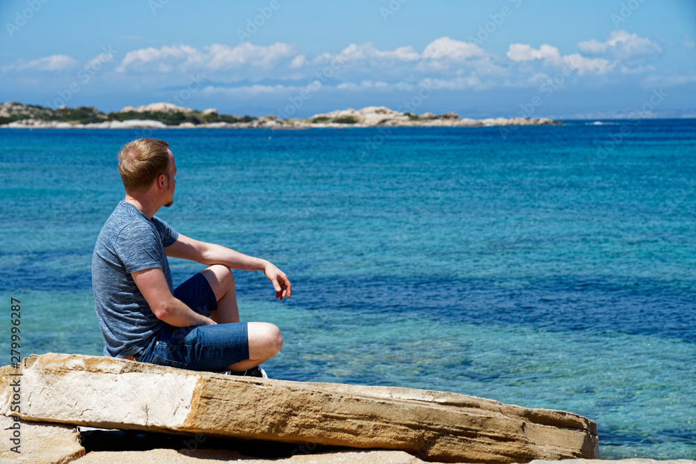 Man sitting on a rock, relaxing and enjoying the beautiful sea view at a beach with a clear blue sky in Sardinia (Italy)