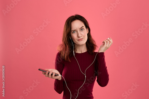 Portrait of a pretty brunette girl holding earphone in one hand and mobile phone in another one standing over pink background. photo