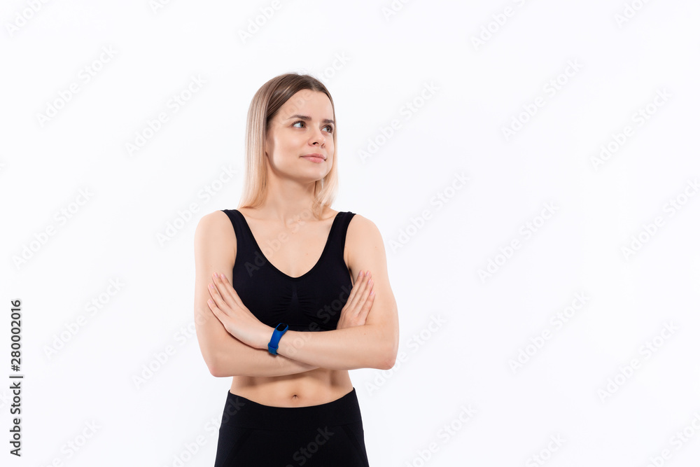 Young sporty blond woman in a black sportswear with smart watches for pulse measuring keeping hands crossed standing over white background.